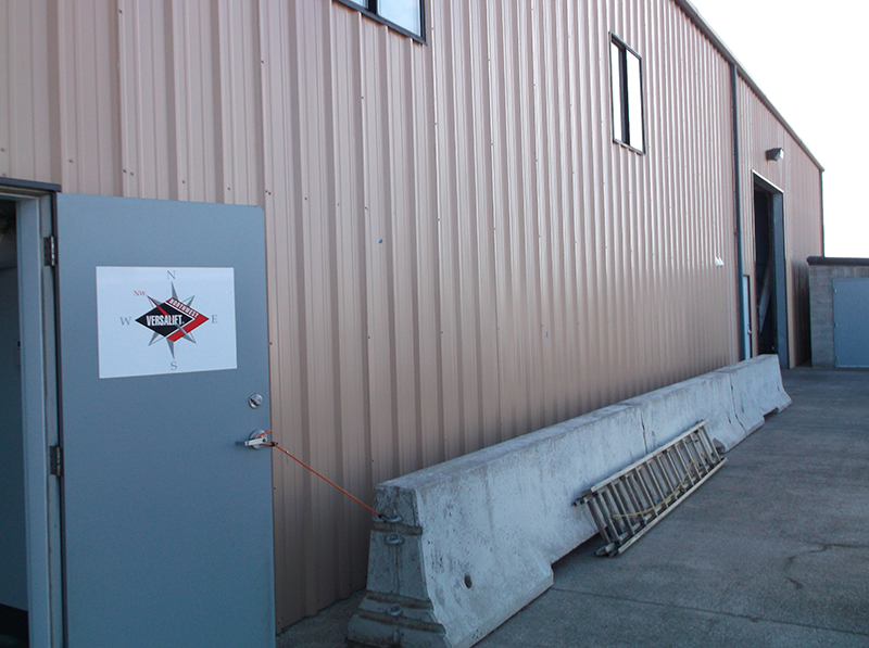 Door open on side of metal paneling industrial building at Brooklane building | Large concrete barriers along wall